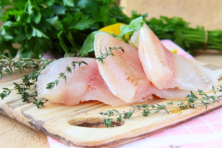 Filés de peixe ao lado de temperos frescos, sobre tábua de madeira