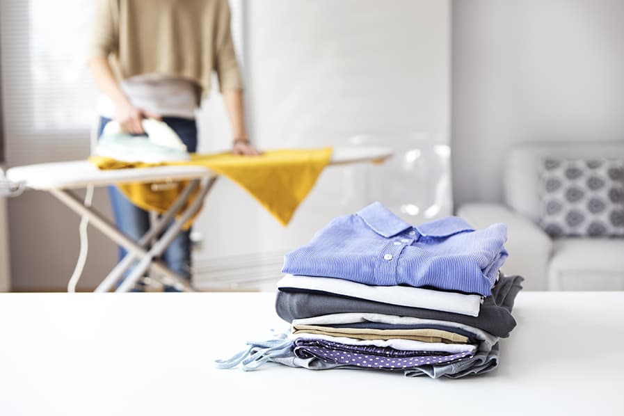 Pilha de roupas dobradas em primeiro plano, ao fundo pessoa passa roupas sobre mesa de passar