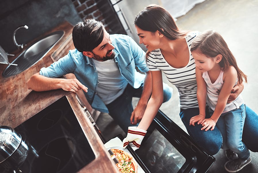 Dois adultos e uma criança em frente à forno com pizza.