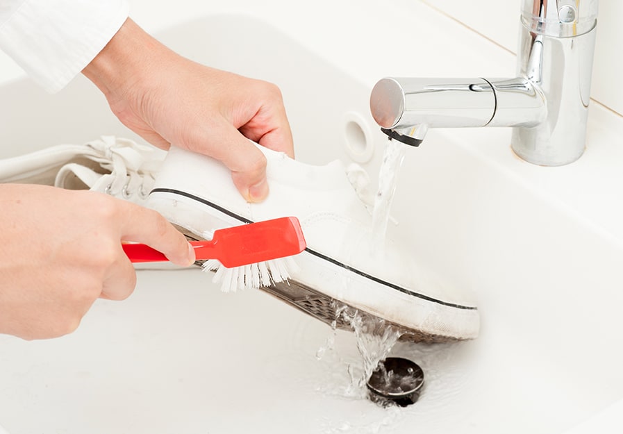  Um tênis branco feito de pano sendo lavado embaixo de uma torneira aberta  e com auxílio de uma escova de limpeza